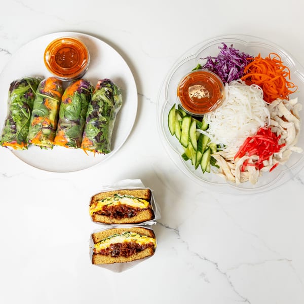 Spring rolls, a noodle salad bowl, and two breakfast sandwiches on a white marble surface.