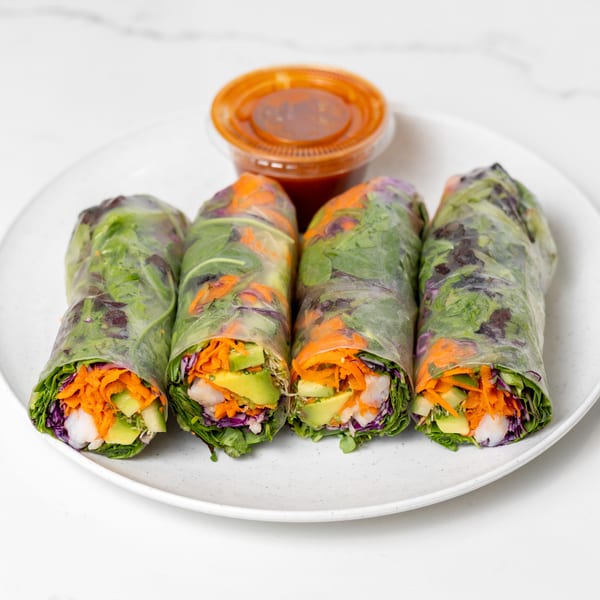 Four fresh spring rolls with shrimp, avocado, carrots, and cabbage, served with a side of sauce.