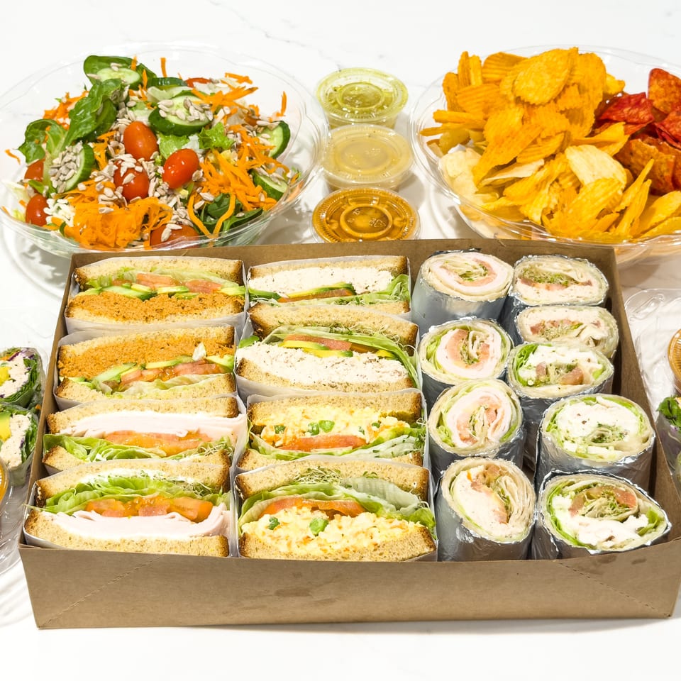 Catering spread featuring sandwiches, wrap pinwheels, salads, vegetable rolls, chips, and individual dressing containers.