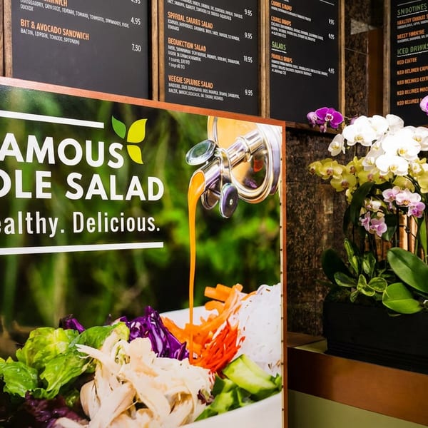Large sign advertising 'The Famous Noodle Salad' with dressing poured over it, next to restaurant menu boards and orchids.