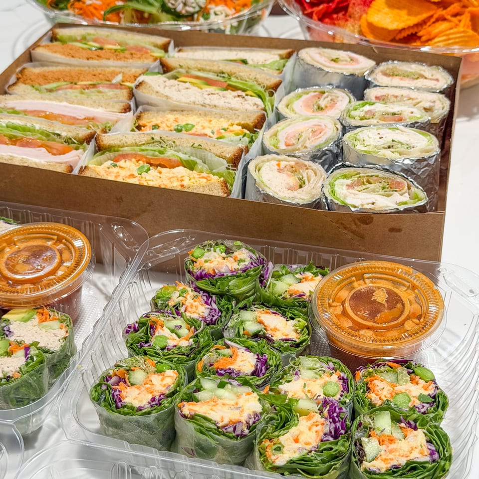 Assorted prepared sandwiches, tortilla wraps, fresh salads, vegetable chips, and dipping sauces arranged in clear takeaway containers.