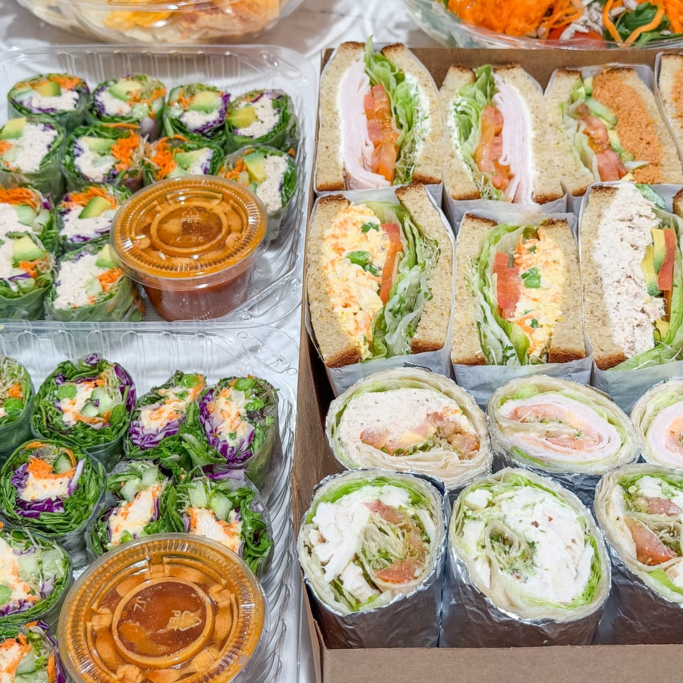 Assorted boxed sandwiches, vegetable wraps, salads, chips, and dipping sauces arranged for restaurant catering.