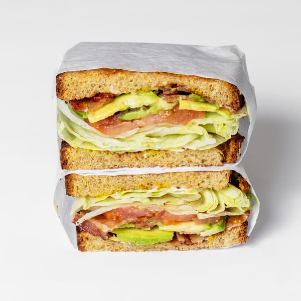 Two halves of a bacon, lettuce, avocado, and tomato sandwich wrapped in white paper.